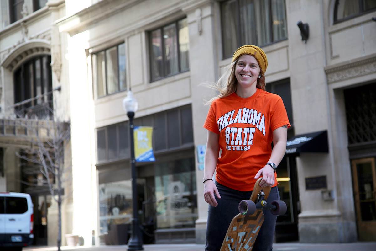 OSU-Tulsa student Rachel Millea in a staged photograph. She is holding a skateboard on a street in downtown Tulsa.